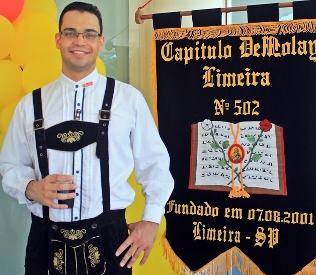 DeMolay com roupa típica alemã em frente à decoração e estandarte do Capítulo Limeira